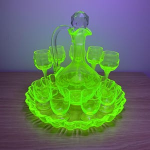 Op de afbeelding: Een fluorescerende geelgroene glazen karafset met een centrale karaf met een gefacetteerde kristallen stop, omringd door zes bijpassende glazen op voet op een geschulpt rond dienblad. Het glaswerk geeft licht onder UV-straling.
