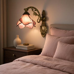 Peut inclure: Une applique murale dotée d'un bras en métal couleur laiton antique orné de volutes complexes et d'un abat-jour en verre en forme de pétale dans une teinte rose douce qui diffuse une lumière chaude dirigée vers le bas au-dessus du lit.
