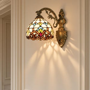 May include: A wall-mounted lamp featuring a dome-shaped stained glass shade with a geometric pattern in cream, blue, yellow, and red. The fixture has an ornate, antique-finished bronze metal arm and a decorative backplate with intricate scrollwork.