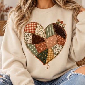 Op de afbeelding: Een beige sweater met ronde hals en een grote hartvormige afbeelding op de borst. Het hart is samengesteld uit diverse patchwork-stijl stoffen met patronen zoals vichy, bloemen, stippen en sterren, afgewerkt met geborduurde madeliefjes.