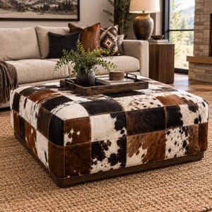 Peut inclure: Un grand pouf carré recouvert d'un patchwork de cuir de vachette présentant un mélange de motifs tachetés blancs, noirs et marron. Le pouf possède une base en bois foncé à profil bas et repose sur un tapis tissé texturé dans un salon.
