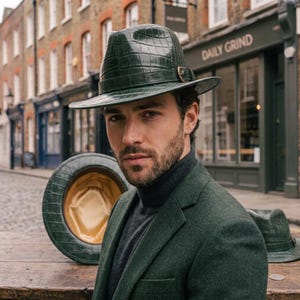 Puede incluir: Un sombrero fedora de color verde oscuro con una textura brillante de piel de cocodrilo, que presenta una banda a juego y una hebilla metálica. El interior del sombrero está forrado con una tela lisa de color dorado. Tiene una forma estructurada.