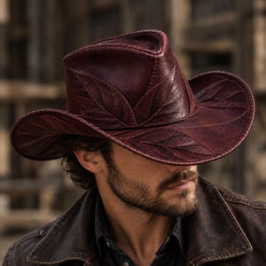 May include: A deep burgundy leather cowboy hat featuring intricate embossed leaf patterns across the crown and the wide, curved brim. The hat displays visible stitching along the edges of the leaf designs and a structured, pinched crown shape.