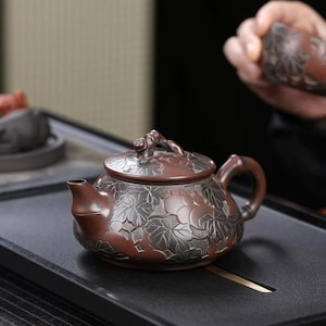 Peut inclure: Une théière en argile brun rougeâtre foncé ornée d'un motif complexe en relief de feuilles et de fruits argentés sur tout le corps arrondi et le couvercle. Elle possède une anse courbée, un bec court et un petit bouton texturé sur le couvercle.