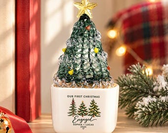 Peut inclure: Un sapin de Noël en verre avec une étoile dorée au sommet et des accents en verre coloré. L'arbre repose dans un pot en céramique blanc carré portant l'inscription "OUR FIRST CHRISTMAS Engaged SOPHIA + LUCAS 2026" et trois sapins imprimés.