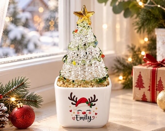 Könnte beinhalten: Ein Weihnachtsbaum aus klarem Glas mit grünen Akzenten und einem gelben Stern als Spitze, in einem weißen, quadratischen Keramiktopf. Der Topf zeigt eine Grafik eines Rentiers mit Weihnachtsmütze und den Namen Emily in schwarzer Schrift.