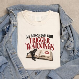 Può includere: Una t-shirt color crema con il testo nero e rosso "MY BOOKS COME WITH TRIGGER WARNINGS" sopra un'illustrazione di un libro aperto con un bordo in pizzo nero e un segno di bacio con rossetto rosso sulla pagina, sotto una giacca di jeans blu.