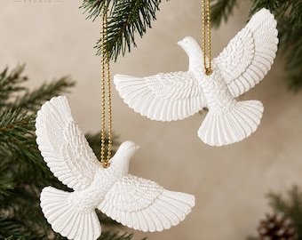 Op de afbeelding: Twee witte duif-ornamenten met gedetailleerde vedertexturen, elk opgehangen aan een goudkleurige kralenketting. De vogels zijn afgebeeld in vlucht met gespreide vleugels. De tekst "figurist STUDIO" staat in de linkerbovenhoek.