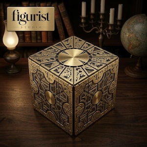 May include: A metallic gold-toned puzzle box featuring intricate, raised geometric and filigree patterns on each face. The cube has a smooth circular center plate on the top and side panels, with deep black recessed areas between the ornate gold designs.