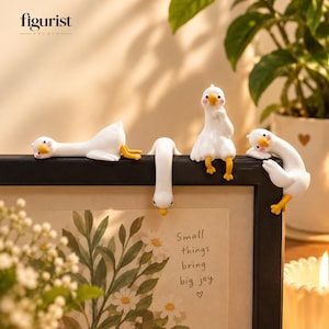 Puede incluir: Cuatro figuras de gansos blancos con picos y patas amarillas están posadas en el borde superior de un marco de fotos negro. El marco contiene un estampado de margaritas y el texto "Small things bring big joy" con un pequeño corazón debajo.