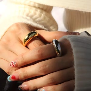 May include: A polished gold-tone dome ring and a polished silver-tone dome ring. Both rings feature a smooth, rounded, thick band design with a high-shine reflective finish, worn on separate fingers to showcase the contrasting metallic colours.
