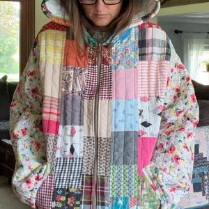 May include: A zip-up hooded jacket constructed from a patchwork of various fabric squares featuring diverse patterns including floral, plaid, gingham, and solid colors in shades of red, blue, yellow, pink, green, and black with a white interior lining.
