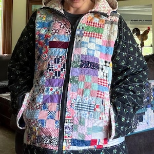 May include: A hooded zip-up jacket featuring a patchwork front panel made of various colorful fabric squares in shades of pink, blue, red, and purple. The sleeves and bottom hem are made of black quilted fabric with a small floral pattern.