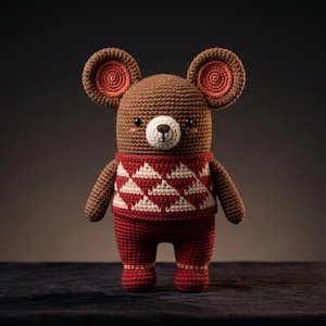 May include: A brown crocheted bear toy with large round ears featuring inner coral circles. The bear wears a red jumper with a white diamond pattern and matching red trousers. It has a white muzzle, a dark brown nose, and small black eyes.
