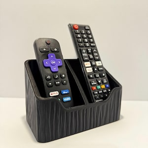May include: A black remote control holder with a vertical ribbed texture on the exterior. The holder features two separate compartments, each containing a black remote control with various buttons, including Netflix, Vudu, and Disney+ logos.