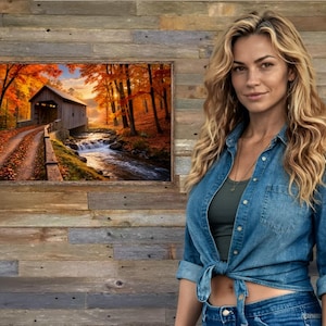Op de afbeelding: Een rechthoekige wandkunstprint met een landschap van een houten overdekte brug over een stromende beek, omringd door herfstbomen met levendig oranje en geel gebladerte, geplaatst tegen een achtergrond van een rustieke houten wand.