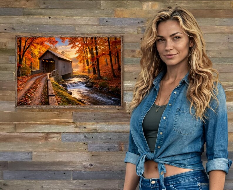 Op de afbeelding: Een rechthoekige wandkunstprint met een landschap van een houten overdekte brug over een stromende beek, omringd door herfstbomen met levendig oranje en geel gebladerte, geplaatst tegen een achtergrond van een rustieke houten wand.