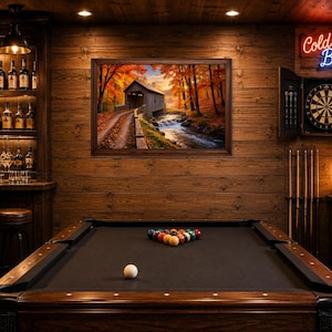 Op de afbeelding: Een speelkamer met een donkerhouten pooltafel, een dartbord aan de muur met een neon Cold Beer-bord, een bar met planken vol glazen flessen en een ingelijst landschapsschilderij van een overdekte brug in een herfstachtig landschap.