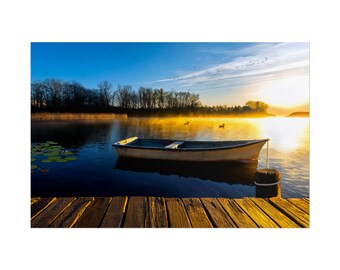 May include: A wooden rowboat floats on a calm lake at sunrise, with a wooden dock in the foreground. The water reflects the golden light of the sun and the blue sky, while mist hangs over the distant treeline and two ducks swim nearby.