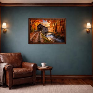 Op de afbeelding: Een ingelijste landschapsafdruk met een overdekte brug over een beekje, omringd door herfstbladeren met oranje en rode bomen. Het kunstwerk hangt aan een donkerblauwe muur tussen twee houten boekenplanken, een leren fauteuil en een bijzettafel.