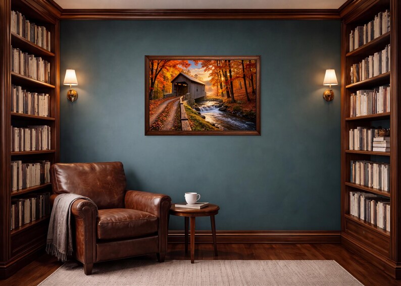 Op de afbeelding: Een ingelijste landschapsafdruk met een overdekte brug over een beekje, omringd door herfstbladeren met oranje en rode bomen. Het kunstwerk hangt aan een donkerblauwe muur tussen twee houten boekenplanken, een leren fauteuil en een bijzettafel.