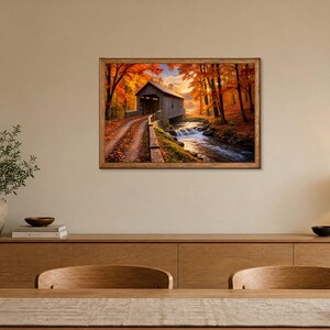 Op de afbeelding: Een rechthoekige kunstdruk in een houten lijst met een overdekte brug over een stromende beek, omringd door levendige herfstbomen met oranje en geel gebladerte, opgehangen aan een muur boven een houten dressoir.