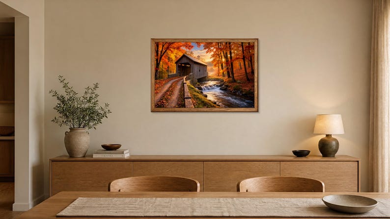 Op de afbeelding: Een rechthoekige kunstdruk in een houten lijst met een overdekte brug over een stromende beek, omringd door levendige herfstbomen met oranje en geel gebladerte, opgehangen aan een muur boven een houten dressoir.