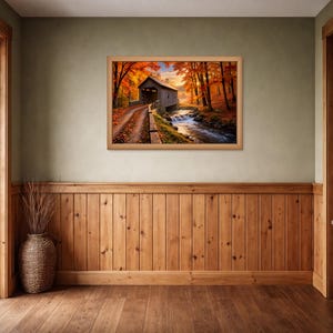 Op de afbeelding: Een rechthoekige ingelijste kunstdruk met een overdekte houten brug over een stromende beek, omringd door herfstbomen met levendig oranje en rood gebladerte, opgehangen aan een lichtgroene muur met houten lambrisering en afwerking.