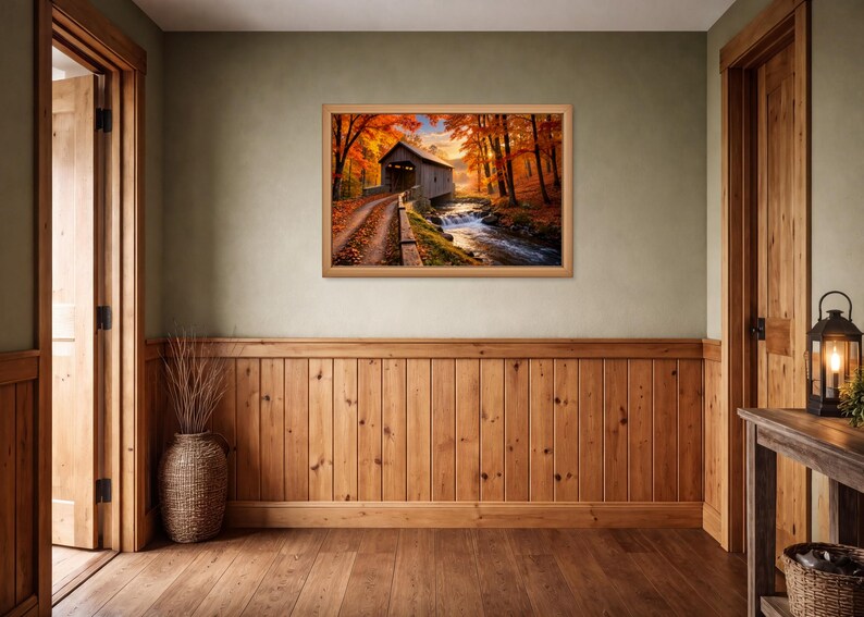 Op de afbeelding: Een rechthoekige ingelijste kunstdruk met een overdekte houten brug over een stromende beek, omringd door herfstbomen met levendig oranje en rood gebladerte, opgehangen aan een lichtgroene muur met houten lambrisering en afwerking.