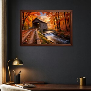 Op de afbeelding: Een ingelijste landschapsmuurprint met een overdekte houten brug over een stromende beek, omringd door herfstbomen met levendig oranje en rood gebladerte onder een zachte zonsonderganglucht, weergegeven in een donker interieur.