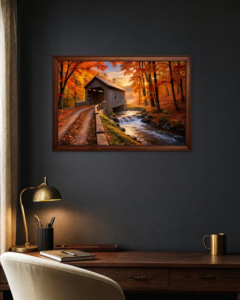Op de afbeelding: Een ingelijste landschapsmuurprint met een overdekte houten brug over een stromende beek, omringd door herfstbomen met levendig oranje en rood gebladerte onder een zachte zonsonderganglucht, weergegeven in een donker interieur.