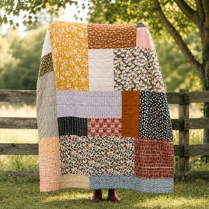 May include: A patchwork quilt featuring a variety of fabric patterns including mustard yellow floral, solid rust orange, black and white daisies, small geometric prints, and gingham checks. The rectangular textile is stitched with a grid pattern.