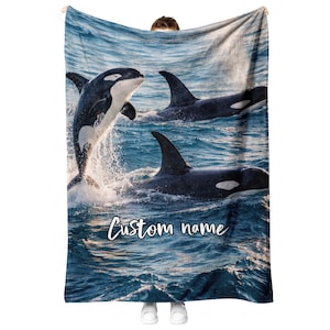 Puede incluir: Una manta rectangular de forro polar con un estampado fotográfico de tres orcas nadando y saltando en aguas oceánicas de color azul profundo. La manta muestra el texto Custom name en una fuente cursiva blanca cerca de la parte inferior.