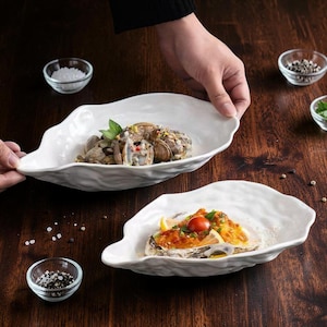 Puede incluir: Dos platos de servicio de cerámica blanca con forma de concha de ostra y bordes irregulares y ondulados. Un plato contiene una porción de almejas cocidas, mientras que el otro contiene una ostra cubierta con un tomate rojo, hierbas y una salsa amarilla.