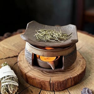 Puede incluir: Un quemador de aceite de cerámica marrón oscuro con exterior texturizado y una bandeja superior cuadrada que contiene agujas de romero seco. Una pequeña vela de té encendida se encuentra en la base. Al lado hay un manojo de romero atado con hilo blanco.