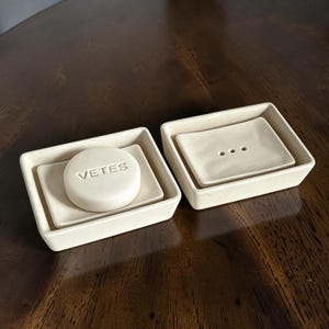May include: Two rectangular off-white ceramic soap dishes with a speckled finish. One dish holds a round off-white soap bar embossed with the text VETES. The other dish is empty and features three small drainage holes in the centre of its base.