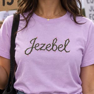 May include: A light purple short-sleeved t-shirt featuring the word Jezebel embroidered in dark green cursive script, with small red thorn-like accents along the letters. The shirt is paired with blue denim jeans and a black shoulder bag.