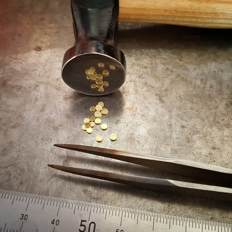 A black metal hammer with a wooden handle, a pair of silver tweezers, and a ruler with markings from 30 to 60. There are small gold metal pieces scattered on a gray metal surface.