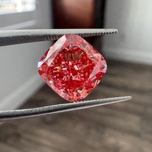 May include: A cushion-cut gemstone with a vibrant, saturated red hue and intricate internal facets that reflect light. The stone is held by a pair of metal tweezers, showcasing its symmetrical shape and deep, brilliant crimson color profile.