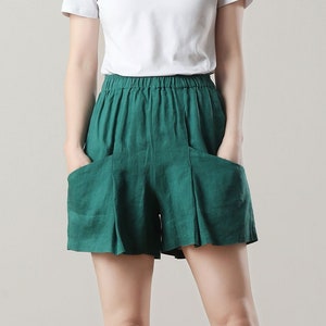 May include: A person wearing a white crew neck t-shirt and green linen shorts with pockets. The shorts have a pleated design. The person is wearing black platform sandals. The background is a neutral color.