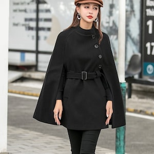 May include: A black cape coat with a belt and buttons. The coat has a high neck and long sleeves. The model is wearing black pants and black boots.
