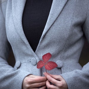 May include: A light gray coat with a black top underneath. A red leaf is held in front of the coat. The coat has three buttons and a classic design. The image has a soft focus.