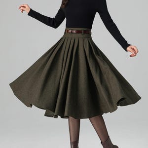 May include: A woman's outfit featuring a black long-sleeve top and a dark green pleated skirt. The skirt is cinched at the waist with a brown belt. The outfit is completed with brown ankle boots.