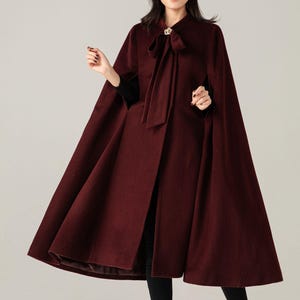 Puede incluir: Un abrigo capa burdeos con un diseño fluido. El abrigo presenta un lazo en el escote y un broche decorativo. La modelo lleva un sombrero negro con velo y botas negras.