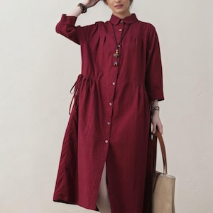 May include: A long-sleeved, button-down dress in a deep red hue. The dress features a collared neckline, a drawstring waist, and a knee-length hem. A beige tote bag with brown leather handles is held in the hand. The person is wearing white shoes.