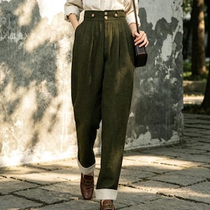 May include: A woman wearing a beige button-down shirt and high-waisted olive green pants. The pants have a pleated design and gold buttons. She is carrying a small black purse and wearing brown loafers.