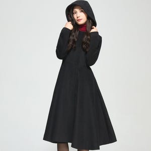 May include: A long black hooded coat with a flared skirt. The coat features a high collar and is worn over a red turtleneck. The model is wearing black tights and black heeled boots. The coat is a classic, elegant design.