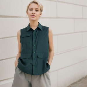 May include: A woman wearing a dark green sleeveless button-up shirt with two pockets and gray shorts.