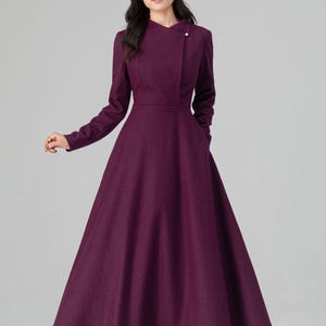 May include: A long, plum-colored dress with a high collar and long sleeves. The dress features a fitted bodice and a flared skirt, with pockets. A single button detail accents the collar. The model is wearing black heels.