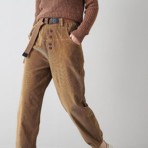 K&ouml;nnte beinhalten: Eine Person tr&auml;gt eine braune Cordhose mit Knopfleiste und einen braunen Strickpullover. Die Hose hat einen G&uuml;rtel mit Schnalle und der Pullover eine Halskette mit Anh&auml;nger.